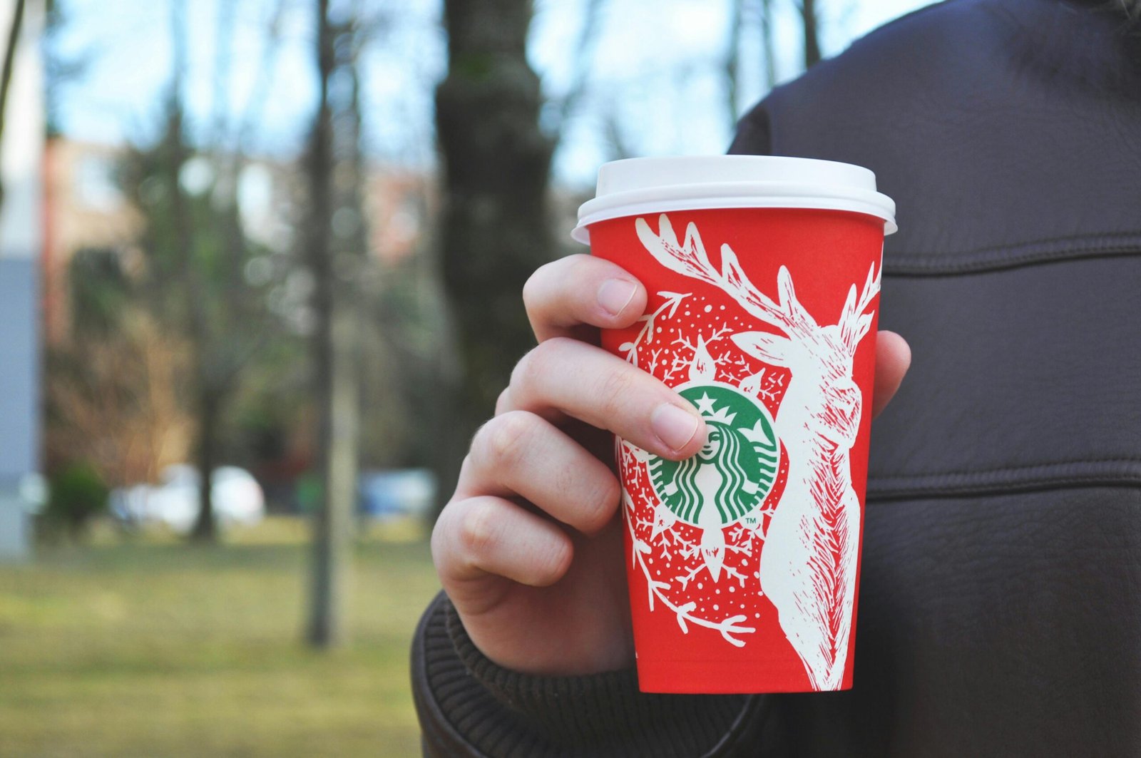 A hand holding a festive red Starbucks cup outdoors in a park setting.